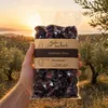 Hand hält Gaia Kallisti Kalamata-Oliven vor Olivenhain Produktbild-Vorschau