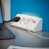 A white power strip with multiple outlets and USB ports is plugged into a laptop on a desk, with a blue wall in the background. Produktbild-Vorschau