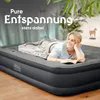 A woman is lying on an inflatable mattress, reading a book. The mattress is set up in a room with plants and a wooden floor. Produktbild-Vorschau