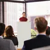A woman is presenting at a podium in front of an audience. Anteprima dell’immagine prodotto