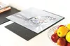 A glass cutting board with a diamond image on it is placed on a black induction hob. A bowl of apples is visible to the right. Produktbild-Vorschau