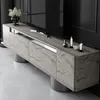A modern TV cabinet with a travertine finish and silver accents, displayed in a dimly lit room with a large black television behind it. Voir les images du produit