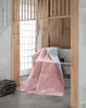 A pink and gray double-faced blanket draped over a chair in a room with wooden accents and a large window. Náhled obrázku produktu