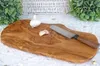 A rustic olive wood cutting board with a knife, garlic bulb, and a garlic clove on a white textured surface, with greenery and a basket in the background. Produktbild-Vorschau