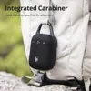 A black portable speaker with a carabiner clip attached to a backpack strap, outdoors. Voir les images du produit