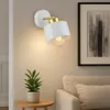 A modern white and gold wall lamp is mounted on a light gray wall above a small wooden side table with a potted plant. A portion of a beige sofa with a pillow is visible on the right. Náhled obrázku produktu