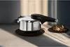 A stainless steel pressure cooker with a black lid and handle sits on a dark wooden trivet, next to a small plate of mushrooms. The scene is lit by soft, directional light, creating shadows on the background and surface. Produktbild-Vorschau