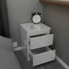 A white bedside table with two open drawers, a black alarm clock, and keys on top, placed next to a gray wall and a bed. Voir les images du produit