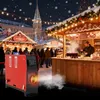 A Christmas market scene at night with a red heater in the foreground and a vendor serving drinks at a stall in the background. Náhled obrázku produktu