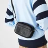 A person wearing a light blue and navy striped sweater is shown from the chest down, with a black Lacoste crossbody bag worn diagonally across their body. Náhľad obrázku produktu