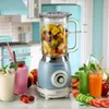A vintage-style blue blender filled with fruit, surrounded by fresh juices and ingredients on a kitchen counter. Anteprima dell’immagine prodotto