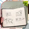 A hand holds open a black mini-notebook with sketches and notes inside, placed on a textured gray surface with a green plant in the background. Náhľad obrázku produktu
