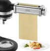 A pasta maker attachment for a stand mixer is shown with a sheet of pasta dough being extruded, and a plate of finished pasta is visible below. Produktbild-Vorschau