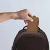 A hand is placing a brown phone case into a brown backpack. Produktbild-Vorschau