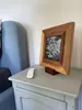 A wooden picture frame stand is placed on a bedside table next to a chair. Produktbild-Vorschau
