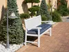 A white garden bench with a dark blue cushion sits on a brick patio next to evergreen trees and a lamppost. Náhled obrázku produktu