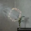 A decorative LED wreath with warm white lights hangs in front of a textured gray wall, with a vase of greenery in the background. Produktbild-Vorschau