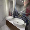 A modern bathroom with two vessel sinks on a wooden vanity, featuring a tropical-themed wallpaper and a large oval mirror. Produktbild-Vorschau