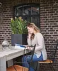 A woman is sitting at a table reading a magazine next to a potted plant. Produktbild-Vorschau