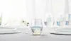 A set of iridescent stemless wine glasses filled with water, arranged on a table with a floral arrangement in the background. Náhled obrázku produktu