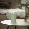 A lit green candle sits on a white table next to a vase with a plant. Podgląd zdjęcia produktu