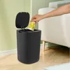 A hand is shown dropping a paper cup with a lemon pattern into a black smart sensor trash can. The trash can has its lid open. The scene is set in a room with a couch and a wall with vertical stripes. Voir les images du produit