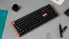 A black mechanical keyboard with orange accents is placed on a gray surface, surrounded by office supplies. Voir les images du produit