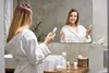 A woman in a bathrobe is holding a cosmetic bottle in front of a mirror in a bathroom. Náhľad obrázku produktu