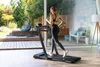 A woman is running on a treadmill in a modern living room with large windows overlooking a garden. Produktbild-Vorschau