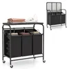 A laundry sorter with an ironing board on top, featuring three black laundry bags and folded clothes and an iron on the ironing board. A smaller inset image shows the same laundry sorter with its lid open. Produktbild-Vorschau
