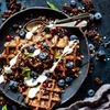 A close-up shot of chocolate waffles topped with blueberries, red currants, granola, and a drizzle of cream, presented on a dark plate. Produktbild-Vorschau