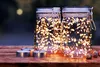 Two glass jars filled with fairy lights sit on a wooden surface, with tea lights and acorns in the foreground. Produktbild-Vorschau