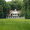 A white house with a thatched roof is visible behind a green mesh fence in a grassy yard. Anteprima dell’immagine prodotto