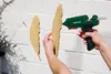 A person is using a green hot glue gun to attach a wooden feather decoration to a white brick wall. Another wooden feather is already attached to the wall. Produktbild-Vorschau