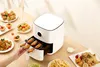 A white air fryer is shown on a wooden table surrounded by various fried foods, with a hand placing a rack of chicken wings into the air fryer. Podgląd zdjęcia produktu