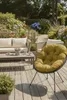 A cozy outdoor seating area with a sofa, coffee table, and a round chair with a yellow cushion, surrounded by lush greenery and flowers. Produktbild-Vorschau
