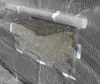 A close-up view of a greenhouse made of transparent plastic sheeting with a mesh reinforcement, showing a rolled-up vent or opening secured with white velcro straps. Anteprima dell’immagine prodotto