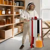 A beekeeper in a protective suit operates a honey extractor, with shelves of honey jars in the background. Produktbild-Vorschau