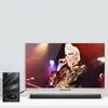 A television displaying a singer performing on stage, with a soundbar and subwoofer positioned below and to the left of the TV. Podgląd zdjęcia produktu