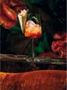 A cocktail glass filled with ice and a reddish-orange drink, garnished with a jalapeño and a small cone of chips, sits on a wooden surface in front of a floral background. Náhled obrázku produktu