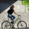 A woman riding an electric bicycle in an urban environment with a concrete wall in the background. Produktbild-Vorschau