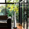 A tall, white and black tower fan is placed on a wooden floor next to a dark brown couch. Large windows with lush green foliage are visible in the background, letting in natural light. Voir les images du produit