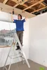A man is standing on a ladder and using a drill to attach a piece of wood to the ceiling. Podgląd zdjęcia produktu