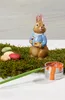 A ceramic Easter bunny figurine dressed in a blue jacket, holding a painter's palette and brush, surrounded by moss and colorful Easter eggs, with a can of orange paint and a paintbrush in the foreground. Produktbild-Vorschau