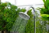 A close-up of an outdoor shower head with water spraying out, set against a backdrop of lush green foliage and a white fence. Produktbild-Vorschau