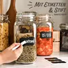 A hand is writing on a label attached to a glass jar filled with pasta. Other jars with food are visible in the background. Podgląd zdjęcia produktu