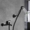 A black faucet and showerhead are mounted on a gray textured wall. Water is spraying from the showerhead. Produktbild-Vorschau