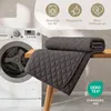 A dark gray quilted blanket is folded and draped over a wooden table next to a washing machine. Icons indicating washability, ease of care, and pet hair resistance are displayed at the top. Náhled obrázku produktu