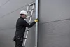 A person wearing a hard hat and work clothes is using an aluminum telescopic ladder against a grey building. Náhľad obrázku produktu