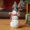 A decorative snowman figurine with a bird and a gift box in the background, set against a red wall and wooden floor. Produktbild-Vorschau